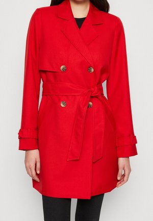 Femme portant un manteau ceinturé rouge vif à double boutonnage avec de gros boutons et des brides aux poignets, par-dessus un pantalon noir.