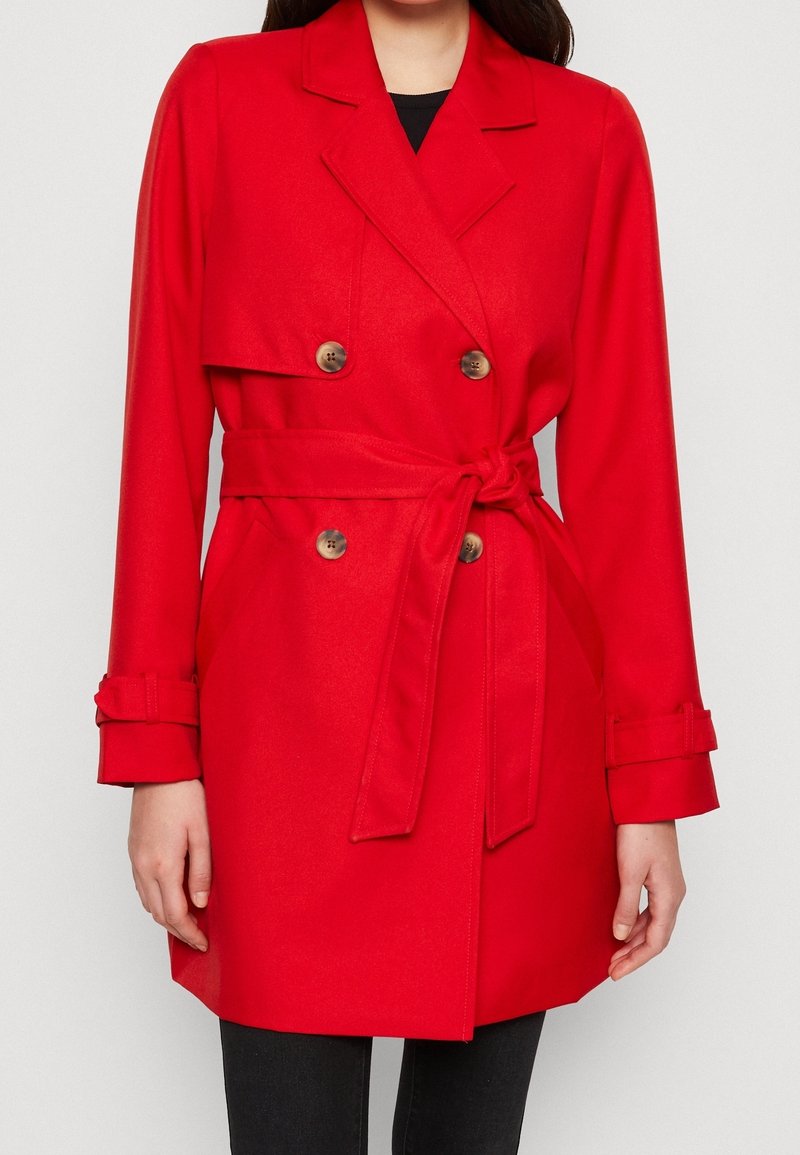 Femme portant un manteau ceinturé rouge vif à double boutonnage avec de gros boutons et des brides aux poignets, par-dessus un pantalon noir.