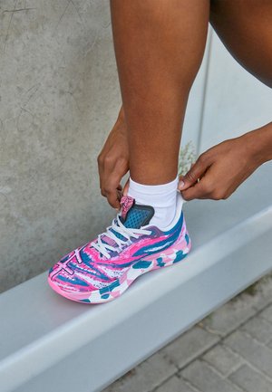 Person tightening white sock over ankle while wearing pink, blue, and white patterned running shoe, resting foot on gray metal rail.