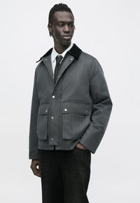 Mand iført en mørkegrå jakke med klaplommer, sort slips, hvid skjorte og sorte bukser, stående med den ene hånd i lommen og kiggende til venstre.