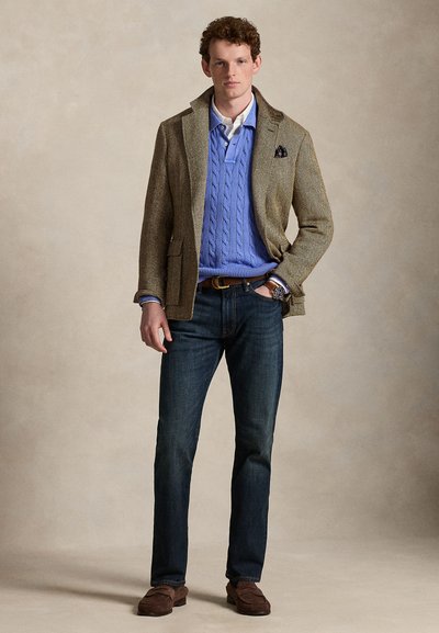 Mladý muž stojí s rukou v vrecku, oblečený v hnedom tweedovom saku, modrý pletený sveter so vzorom cez bielu košeľu s golierom, tmavé rifle a hnedé mokasíny.