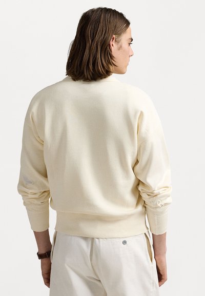 Sweatshirt de culoare crem, cu o croiala lejera, manșete ribbate și guler rotund. Prezintă o textură subtilă și un mic detaliu uzat pe manșon.