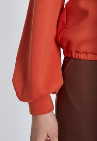 Langärmliges Oberteil aus leuchtendem Korallestoff mit elastischen Bündchen, kombiniert mit einer glatten, braunen, taillierten Unterhose. Strukturierter, leicht dehnbarer Stoff.