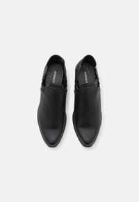 Zapatos negros sin cordones hechos de cuero texturizado, con una punta afilada y sutiles detalles en relieve en la parte superior.