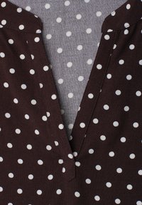 Vêtement marron à col en V avec des pois blancs sur un tissu gris avec un motif similaire de pois blancs.