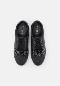 Baskets synthétiques noires avec un logo métallique rond en accent, fermeture à lacets et surface texturée. L'intérieur présente un logo sur la semelle intérieure rembourrée.