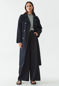 Tume trench coat kahekordsete nupudega ja laia kraega, kombineeritud helesinise sviitriga ja mustade laia säärtega pükstega.