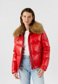 Giacca imbottita rossa con finitura lucida e cappuccio foderato in pelliccia. Presenta una chiusura con zip, tasche laterali e un design aderente. Jeans di denim indossati sotto.