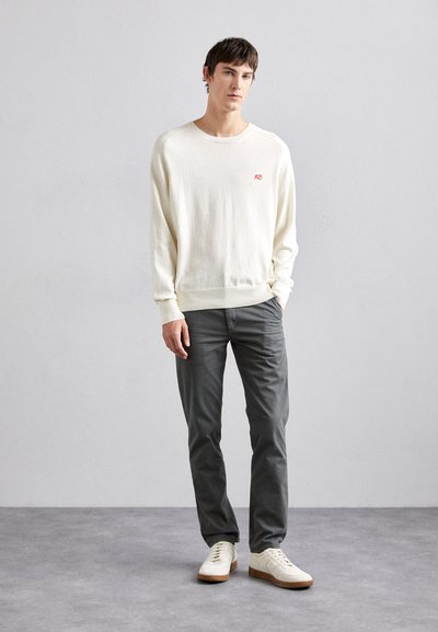 Pulover din tricot de culoare crem deschis, cu mansete cordonate, pantaloni gri și adidași albi cu tălpi bej. Design simplu și branding minim.