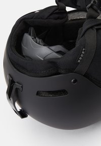 Čierna prilba s matným povrchom, polstrovaným interiérom a nastaviteľným systémom popruhov. Obsahuje ventilačné otvory a bezpečný mechanizmus upevnenia.