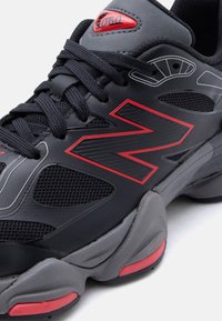 Schwarzer und grauer Sportschuh mit roten Akzenten, schwarzen Schnürsenkeln, Mesh-Einsätzen und einem roten "N"-Logo an der Seite.