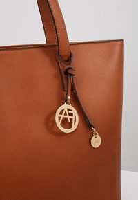 Hnědá kožená kabelka s prošitým popruhem a dvěma kulatými zlatými kovovými logovými přívěsky připevněnými černou zavázanou koženou šňůrkou.