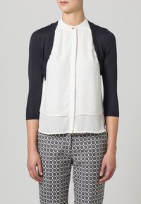 Marinblå 3/4-ärmad cardigan över vit blus, med knäppning och lagerförsedd nederkant. Bärs med mönstrade, högmidjade byxor i mörka färger.