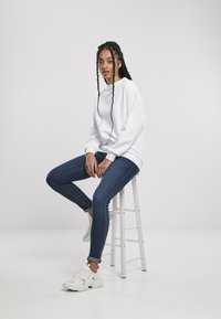 Felpa bianca, jeans skinny blu e sneakers bianchi. Seduto su uno sgabello bianco, con capelli intrecciati e uno sfondo neutro.
