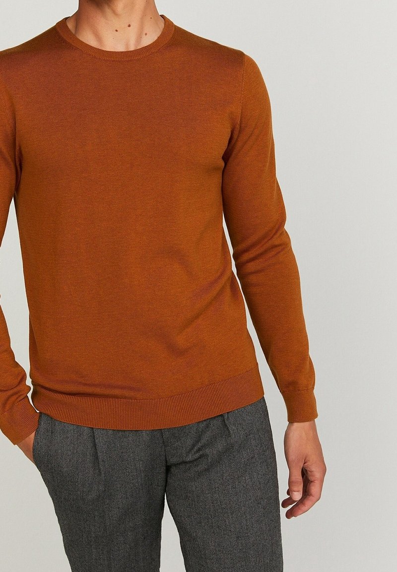 Sweter z okrągłym dekoltem w kolorze spalonej pomarańczy, o prążkowanej fakturze, wąski krój, długie rękawy, zestawiony z szarymi, dopasowanymi spodniami.