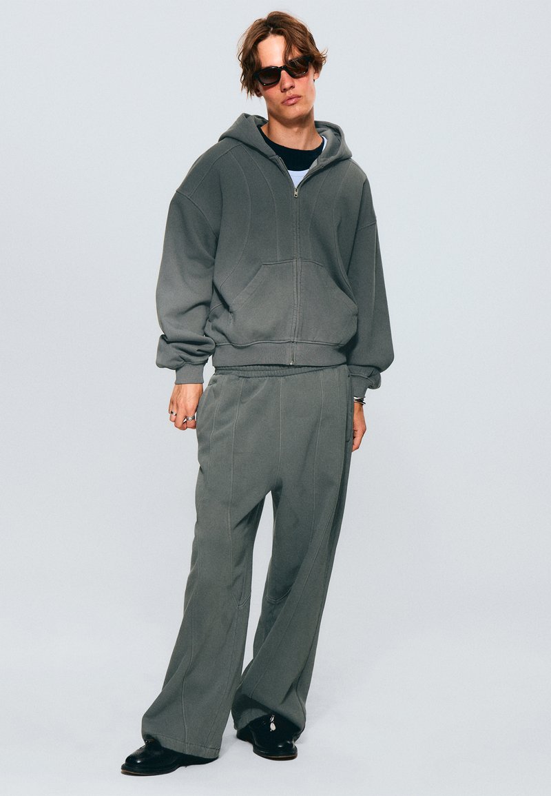 Grauer Hoodie und passende Hose aus weichem Material, mit einem Reißverschluss vorne, geräumigen Taschen und dekorativen Nähten. Getragen mit schwarzen Schuhen.