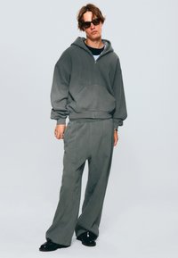 Hanorac oversized gri cu fermoar și buzunar frontal, asortat cu pantaloni largi care au detalii de cusătură laterală. Pantofii negri completează ținuta.