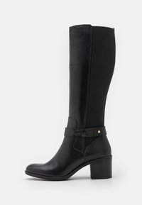Bota de cuero negra hasta la rodilla con panel de tela, tacón bajo, punta redonda y correa decorativa con remache metálico en el lateral.