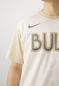 Tricou cu mânecă scurtă în tonuri de crem și bej, având o grafică îndrăzneață "BULL" în auriu, cu contur verde închis și un logo Nike în verde închis.
