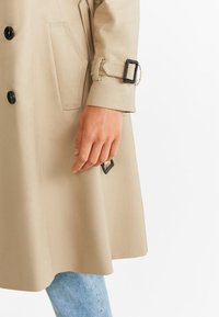 Manche et côté d'un trench-coat beige avec des boutons noirs et une boucle de ceinture, porté sur un jean bleu clair, une main reposant naturellement.