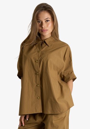 Donna con capelli lunghi che indossa una camicia marrone ampia con bottoni e pantaloni abbinati, in piedi su uno sfondo bianco neutro.