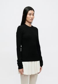 Pulover negru cu dungi și mâneci lungi, cu guler rotund, purtat peste o fustă albă plisată. Textură moale, design strâmt și manșete prelungite.