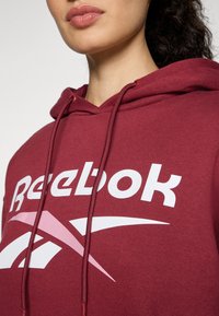 Felpa color amaranto con cappuccio e lacci, grande logo bianco "Reebok", texture liscia e leggero lucido, con un design accentato in rosa sotto il logo.