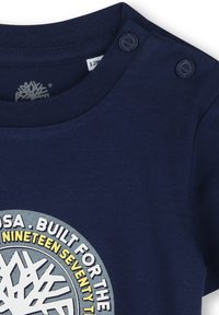 Camiseta de algodón azul marino con escote redondo, detalles de botones a presión en el hombro y un gráfico circular en texto blanco y amarillo con un diseño de árbol.