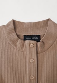 Brun ribbet stoff topp med rund halsutringning, med fire knapper og en svart etikett som leser "ANNA FIELD" inne i kragen.