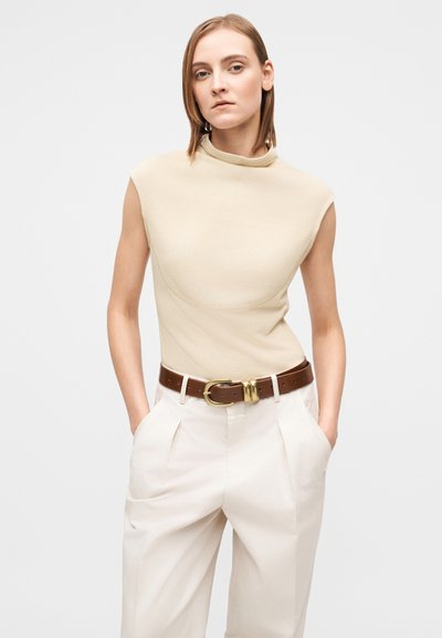 Beige ärmlös topp med hög krage, kombinerad med ljusfärgade plisserade byxor, accentuerade med ett brunt läderbälte med ett guldfärgat spänne.