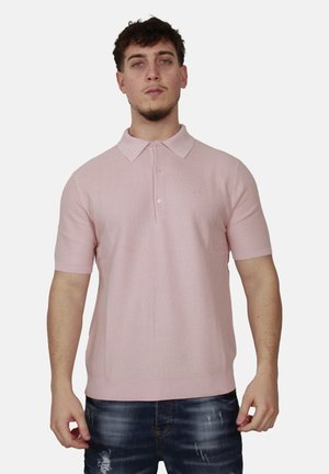 Uomo che indossa una polo a maniche corte rosa chiaro e jeans blu scuro strappati, in piedi contro uno sfondo chiaro e uniforme.