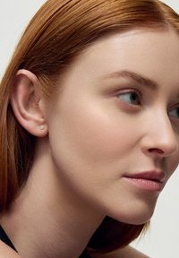 Złote kolczyki wkrętki z motywem kwiatowym; modelka ma gładką skórę oraz proste, średniej długości rude włosy, na jasnym tle.