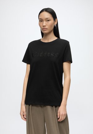 Femme aux longs cheveux noirs porte un t-shirt noir avec le texte « Goddess » et un pantalon beige ample, debout devant un fond blanc uni.
