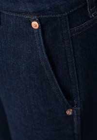 Un jean en denim bleu foncé présente des rivets en cuivre aux poches, une texture lisse et un design classique à cinq poches avec des détails de couture subtils.