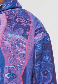 Felpa con cappuccio lavorata a maglia colorata, caratterizzata da motivi astratti in viola, blu e rosso, e un logo Colucci ricamato di colore azzurro chiaro sul petto.