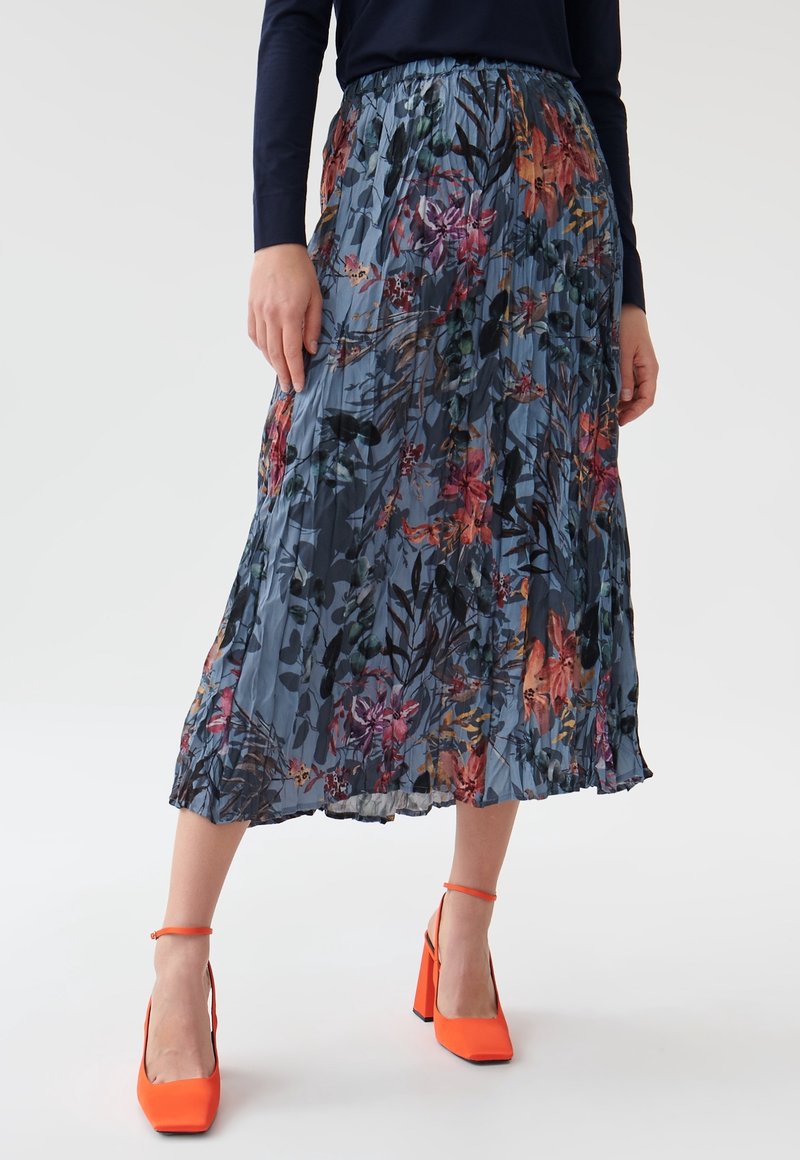 Jupe plissée longueur midi avec base bleue et motif floral multicolore, portée avec un haut bleu marine et des chaussures à talons hauts orange vif.