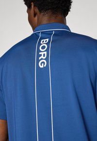 Plava polo majica s bijelim akcentnim linijama i ispisanim "BORG" logotipom okomito na leđima, s klasičnim ovratnikom i kratkim rukavima.