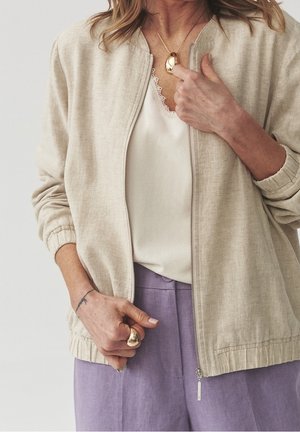 Frau trägt beige Jacke mit Frontreißverschluss, weißes Top mit Spitzenbesatz, lavendelfarbene Hose, hält goldene Anhänger-Halskette und Ring mit sichtbarem Unterarmtattoo.