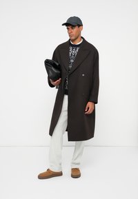 Cappotto lungo marrone con un ampio colletto, indossato sopra un maglione a fantasia, abbinato a pantaloni beige e scarpe marroni. Tiene una borsa nera.