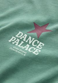 Tricou verde din bumbac, cu o stea maronie mare și textul "DANCE PALACE" în alb, plus "Violet Street Downtown LA" dedesubt.