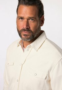 Kermaantuoksuinen nappikauluspaita, jossa on kaksi etutaskua, niittinapit, kaulus ja kankaan kuvioitu pinta, yllään parrallinen mies.