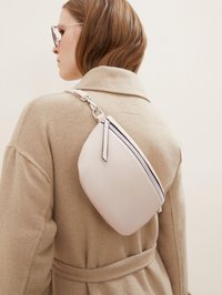 Borsa a tracolla rosa chiaro con superficie texture, chiusura con zip e una cinghia sottile, indossata sopra un cappotto beige. Accenti in metallo argentato.