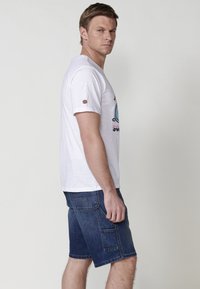 White cotton t-shirt with short sleeves featuring a colorful graphic design on the front. Paired with dark blue denim shorts with pockets.