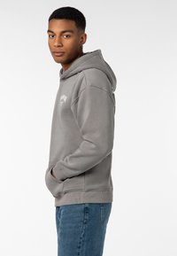 Grauer Pullover-Hoodie mit einer Fronttasche, gerippten Bündchen und einer Kordelzugkapuze. Minimalistisches Logodesign auf der Brust. Getragen mit blauen Jeans.
