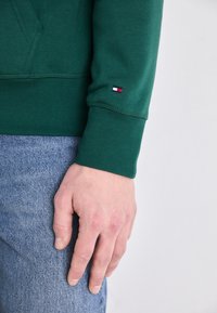 Grüner Baumwollpullover mit einem gerippten Bündchen, versehen mit einem kleinen rot-weiß-blauen Logo am Ärmel. Kombiniert mit Jeans.