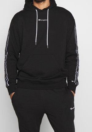 Mann trägt einen schwarzen Champion-Hoodie mit weißen Kordeln und passende schwarze Champion-Jogginghose, Hände in den Taschen.