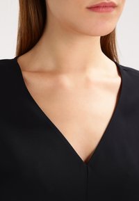 Černý oděv s výstřihem do V, vyrobený z hladké, lehké tkaniny a s minimem švů. Důraz je kladen na výstřih.