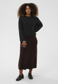 Crni pulover dugih rukava uparen s tamnosmeđom suknjom do koljena. Glatka tekstura; model nosi crne mokasine. Jednostavan i prilagođen dizajn.