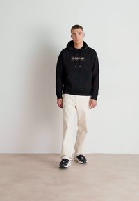 Felpa nera con cappuccio e logo, pantaloni beiges a gamba dritta e sneakers nere, posata su uno sfondo chiaro e semplice.