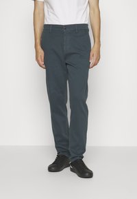 Chinos de algodón azul con un corte recto, cierre de botón, bolsillos frontales y bolsillos laterales, combinados con zapatillas negras y una camiseta blanca.